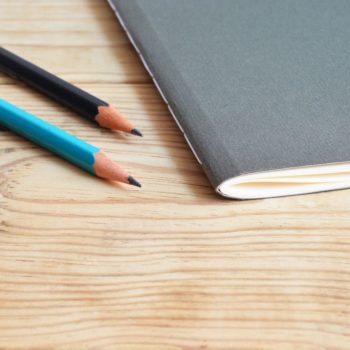 Two pencils and a grey notebook on a light wooden desk