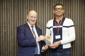 Photo of an older white man with a tall Asian man, standing in front of wooden paneling holding a small trophy